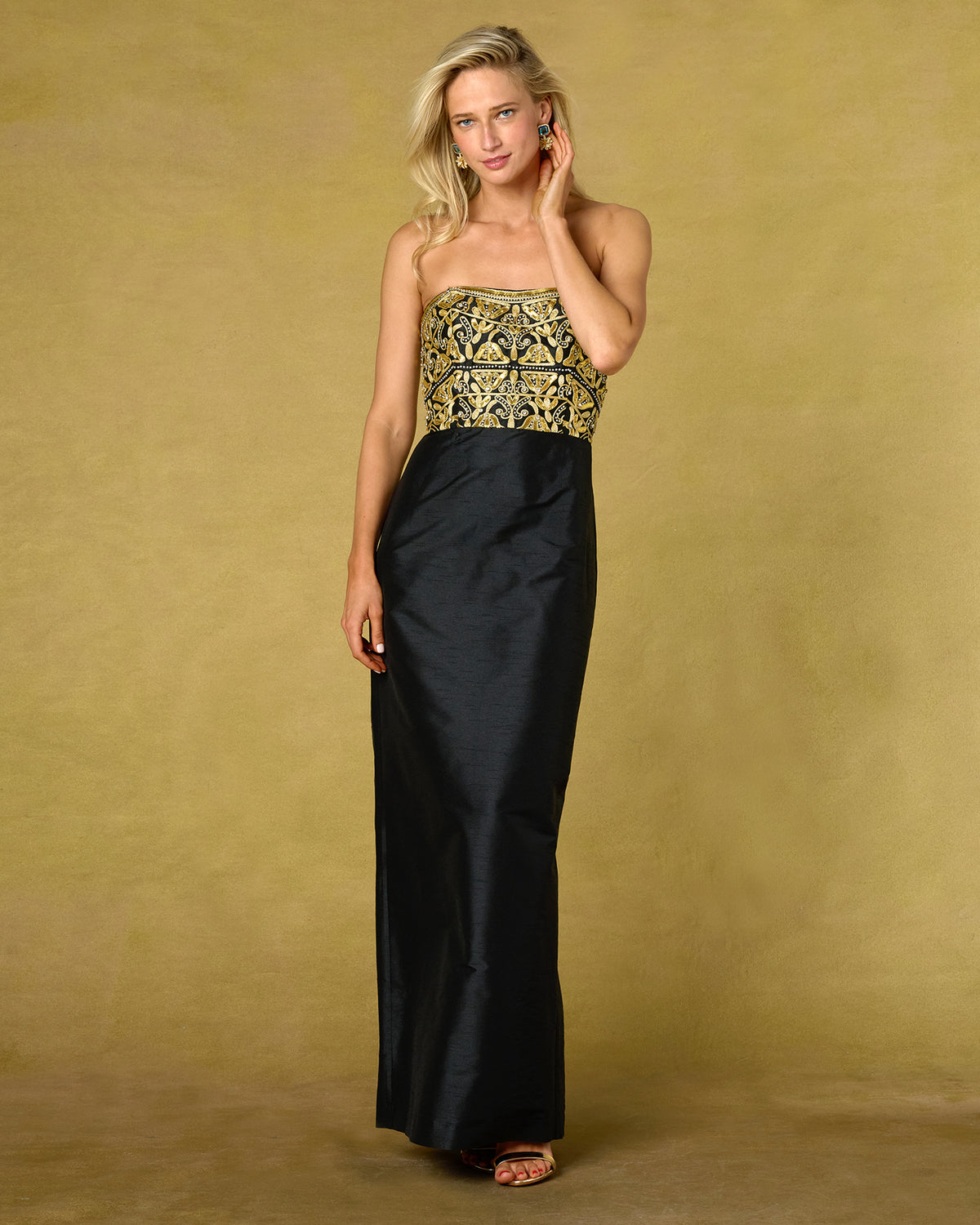 A woman models a strapless maxi dress with a black satin skirt and a gold embroidered bodice, set against a textured gold studio backdrop.