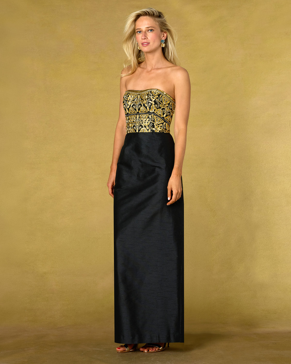 A woman models a strapless formal gown with a black satin skirt and a gold embroidered bodice, set against a textured gold studio backdrop.