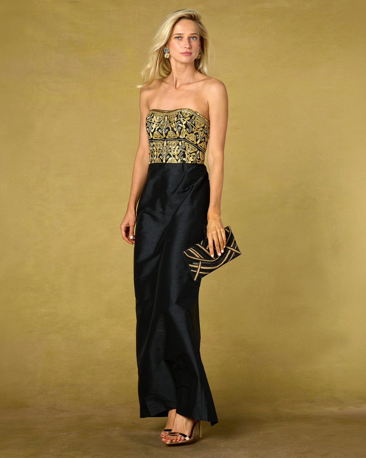 A woman models a strapless black gown with an ornate gold embroidered bodice, holding a matching black and gold clutch against a solid gold studio backdrop.