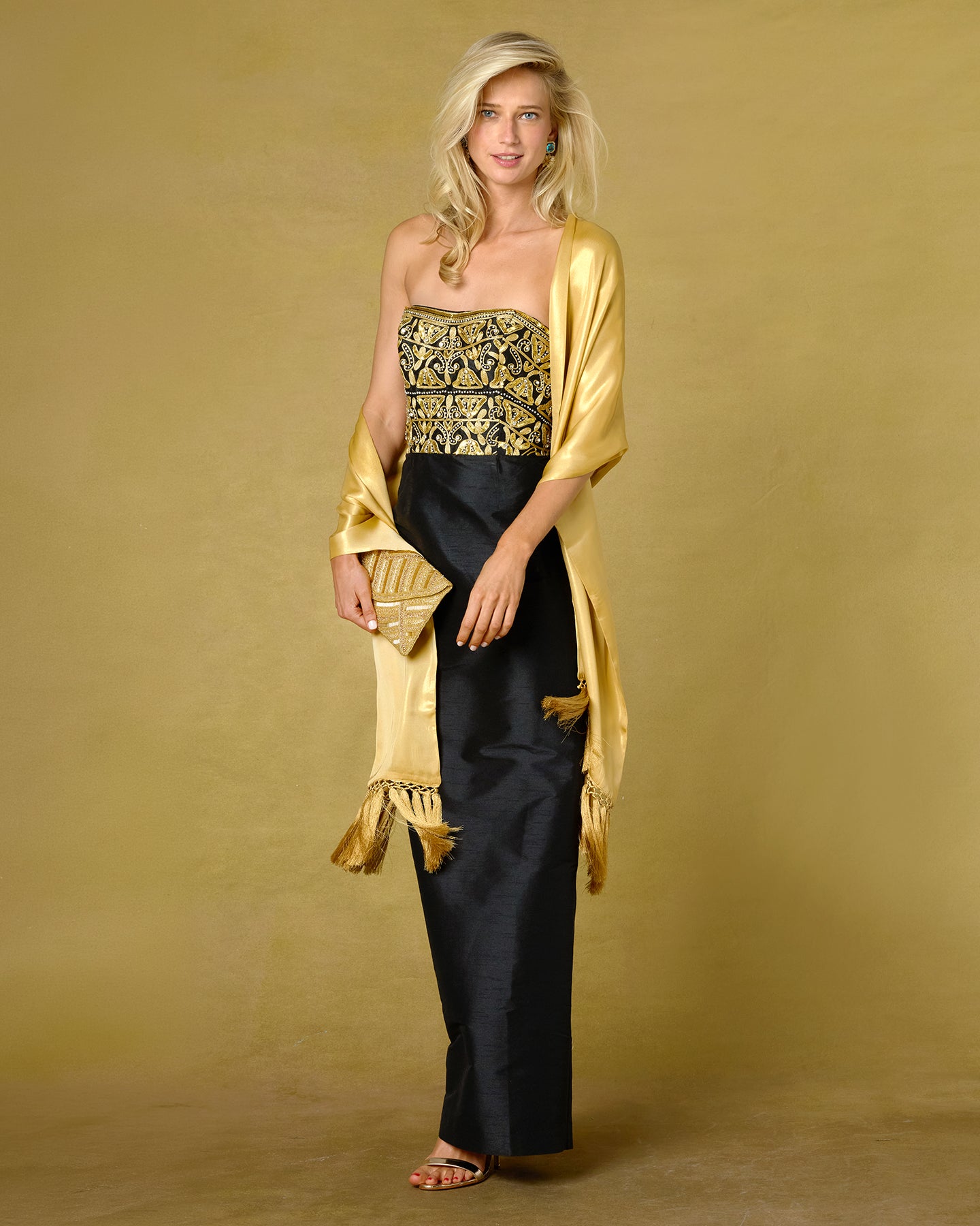 A woman models a strapless black dress with a gold and black patterned bodice, styled with a gold fringed shawl against a textured gold backdrop.