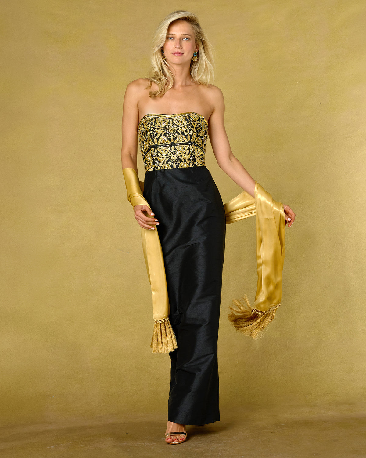 A woman models a strapless black satin gown with a heavily embroidered gold and black bodice, paired with matching long gold gloves and a long gold sash, set against a solid gold backdrop.