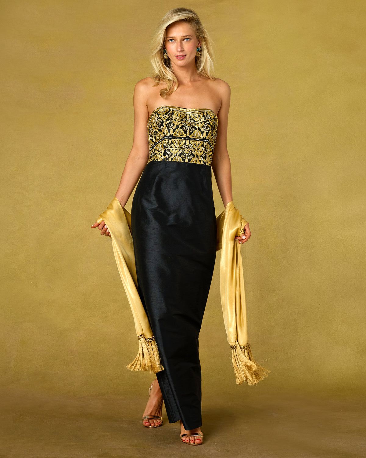 A woman models a strapless black gown with a gold embroidered bodice and holds a gold fringe shawl against a solid gold studio backdrop.