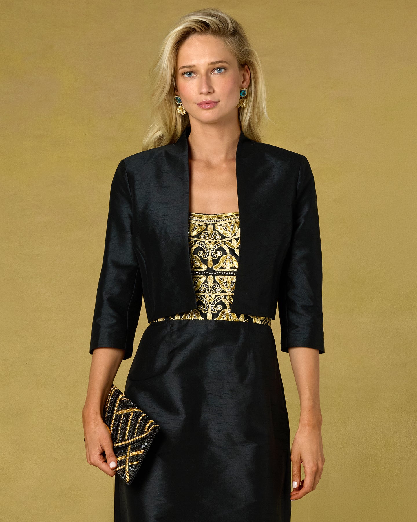 A blonde model wearing a black three-quarter sleeve jacket over a black dress with a gold embroidered bodice, holding a black and gold patterned clutch against a solid gold background.