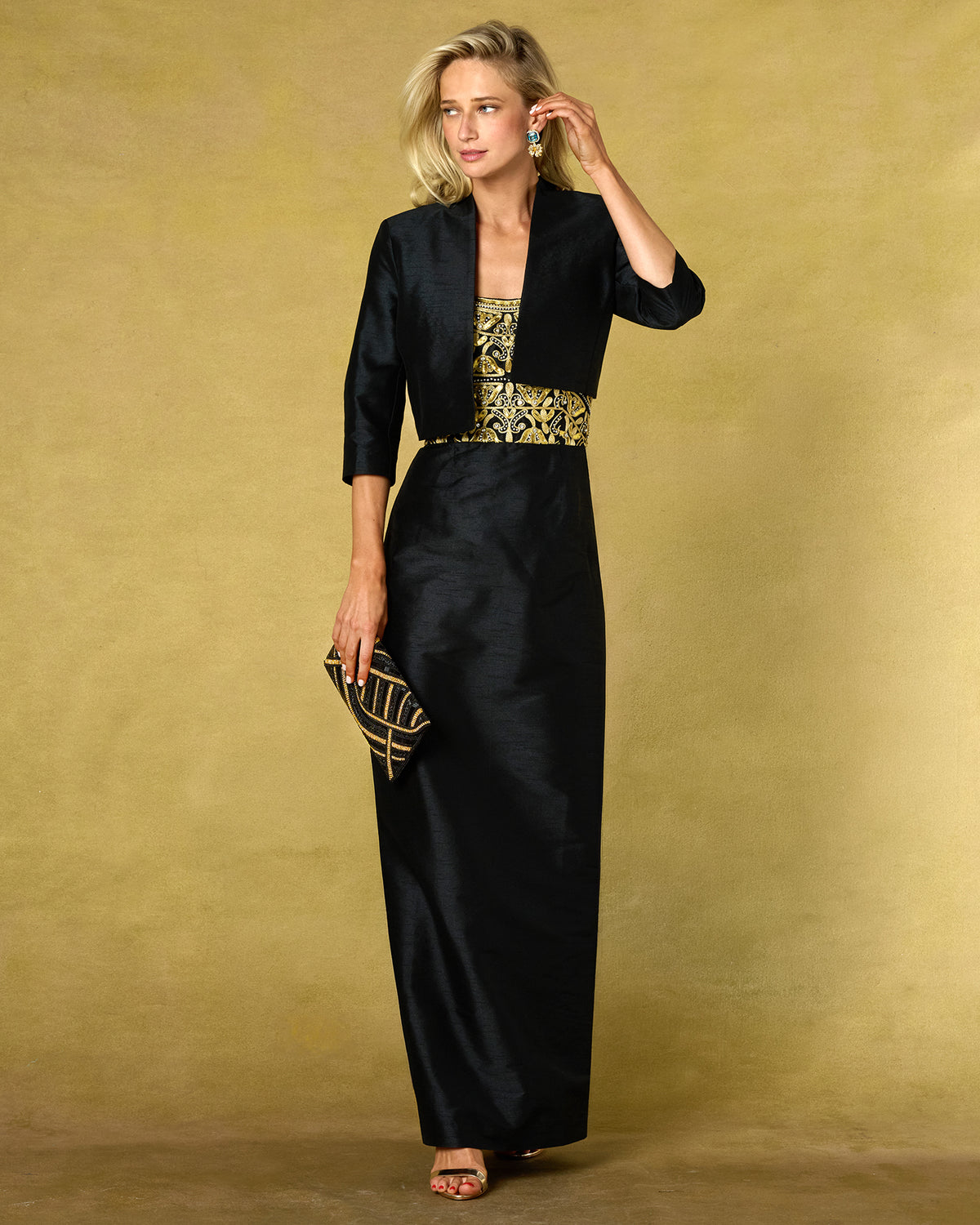 A woman models a black floor-length gown with a matching cropped jacket, featuring gold embroidery on the bodice and a matching clutch, set against a textured gold studio backdrop.