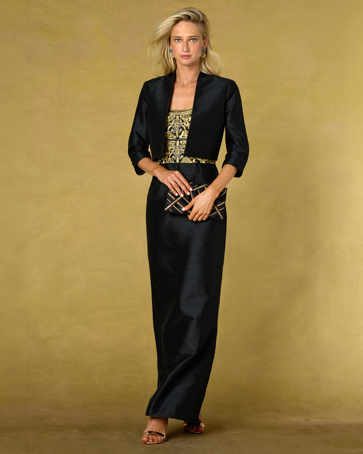 A woman models a black satin-like maxi dress with three-quarter sleeves and a matching jacket-style collar, styled with a gold patterned top underneath and a gold belt, against a textured gold studio backdrop.