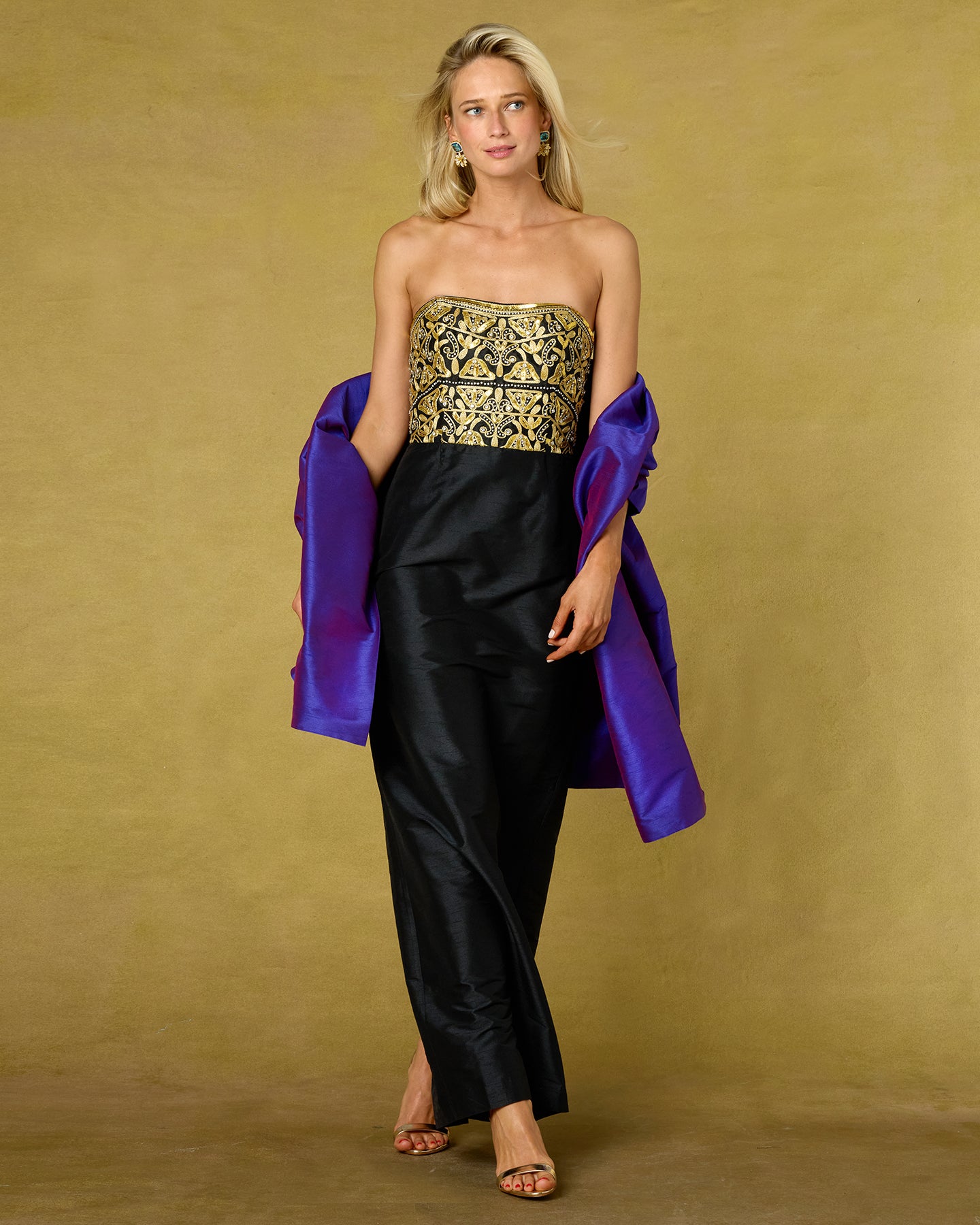 A blonde model stands against a textured mustard-yellow studio backdrop wearing a strapless black gown with an ornate gold embroidered bodice and holding a bright purple satin wrap draped over her shoulders.