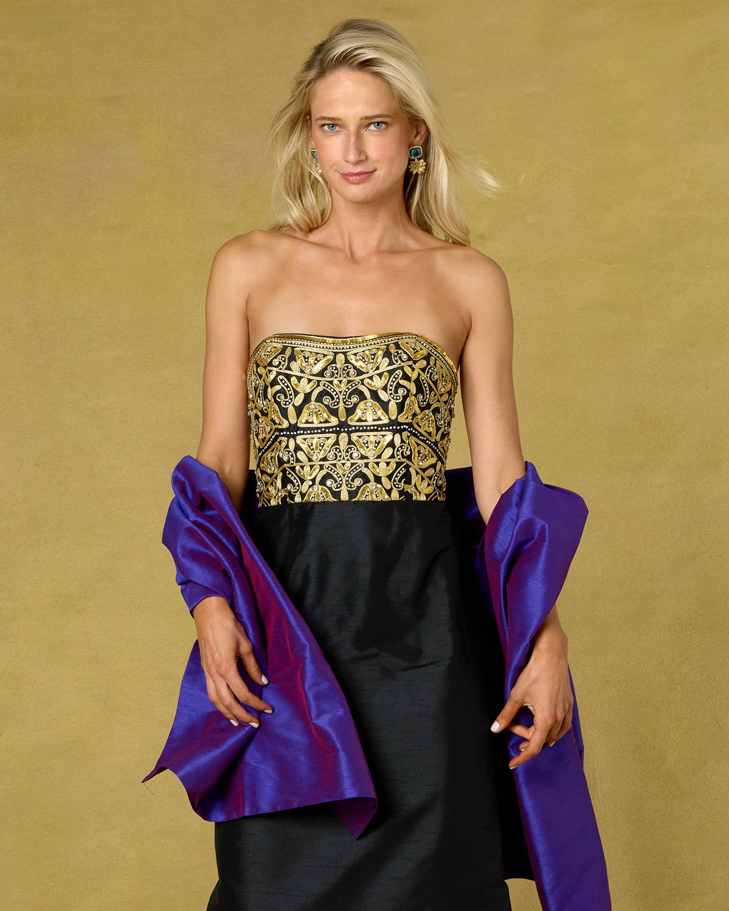 A blonde model wears a strapless black dress with an ornate gold embroidered bodice and holds purple satin sleeves or shawls against a warm gold studio backdrop.