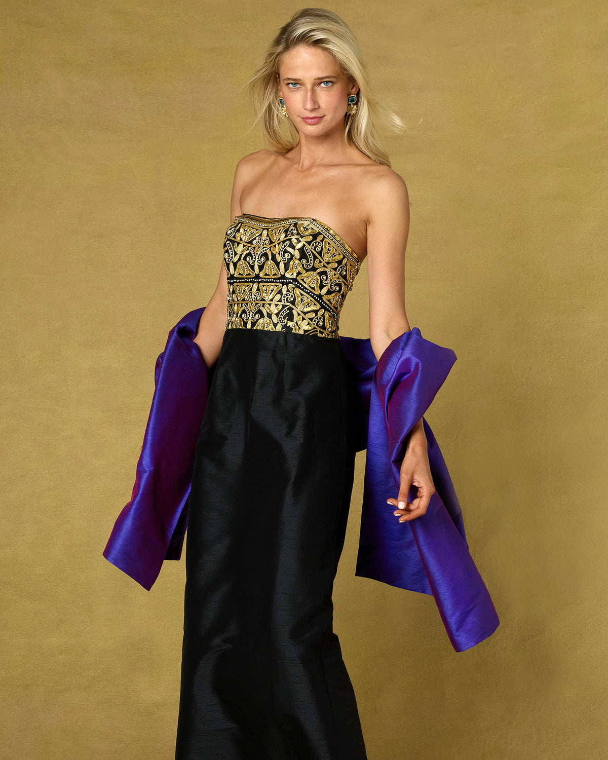 A blonde model wears a strapless black gown with a heavily embroidered gold bodice and holds a bright purple satin taffetta dupioni shawl against a textured gold studio backdrop.