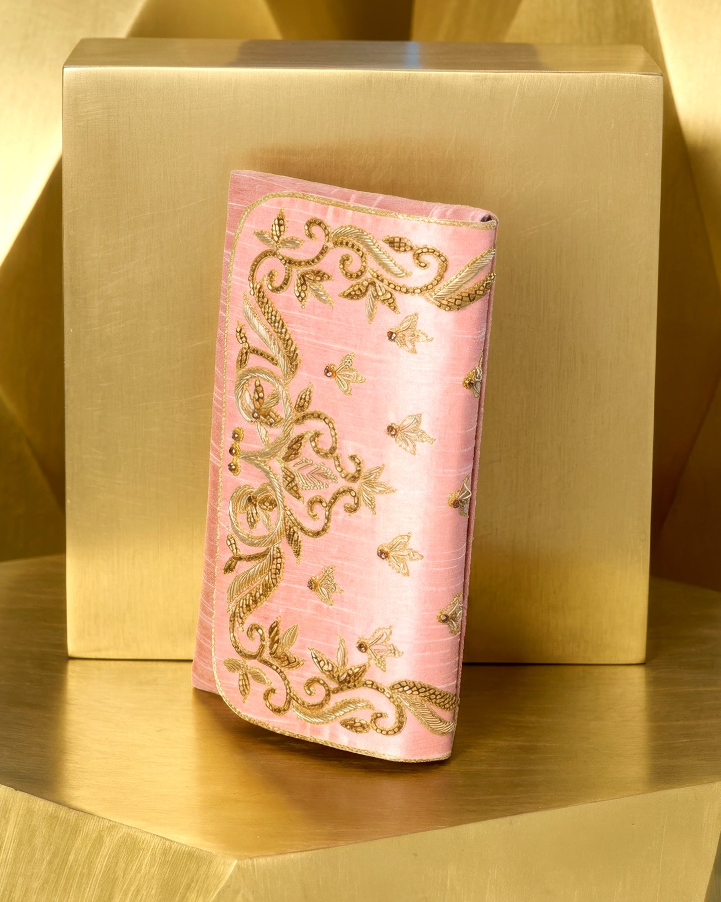Pink embroidered clutch bag on a gold surface with a gold box in the background