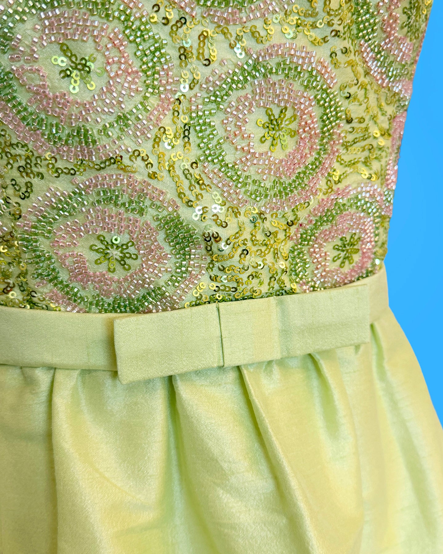 Close-up of a light yellow-green dress featuring a heavily embellished bodice with pink and green sequins and beadwork, paired with a matching satin waistband and pleated skirt.