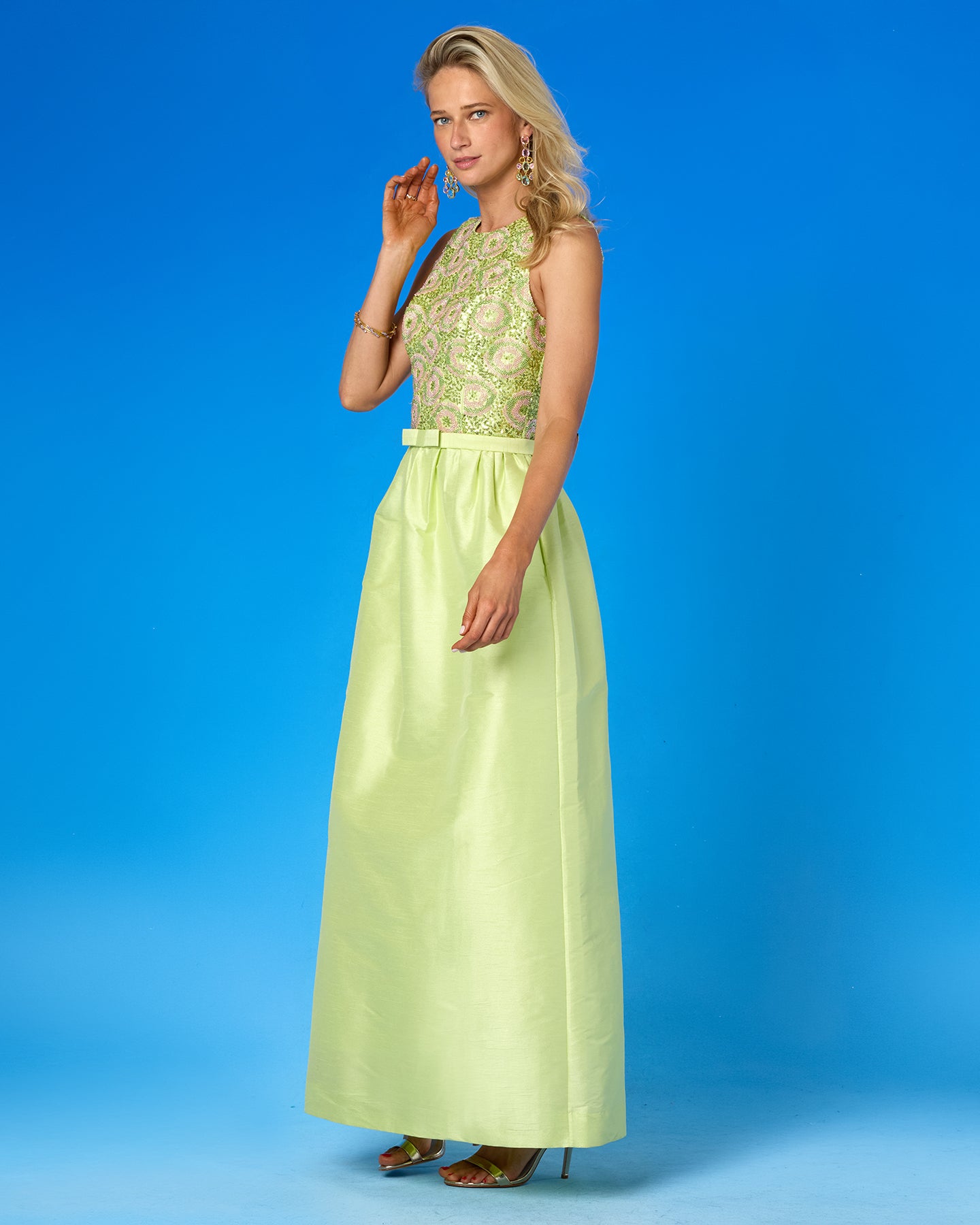 A woman modeling a sleeveless formal gown with a light green satin skirt and a heavily embroidered bodice in matching green and gold tones, set against a solid blue studio background.
