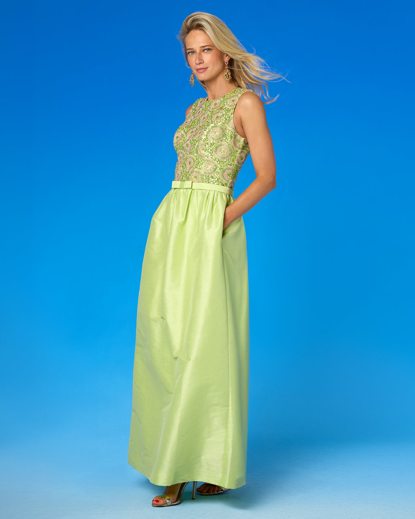A woman modeling a sleeveless maxi dress with a light green satin skirt and a heavily embroidered bodice in green, pink, and gold, set against a solid blue studio background.