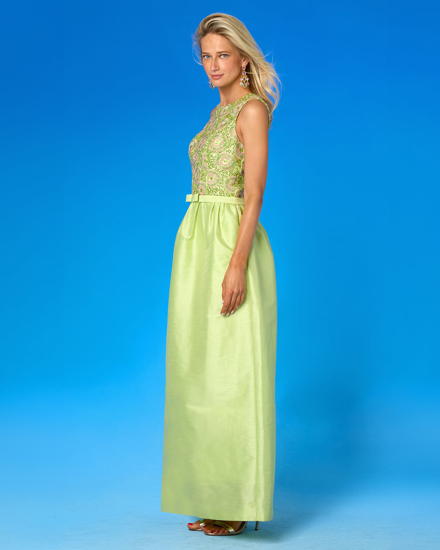 A woman modeling a sleeveless formal gown with a light lime green satin skirt and a heavily embroidered bodice in matching green and gold tones, posed against a solid blue studio background.