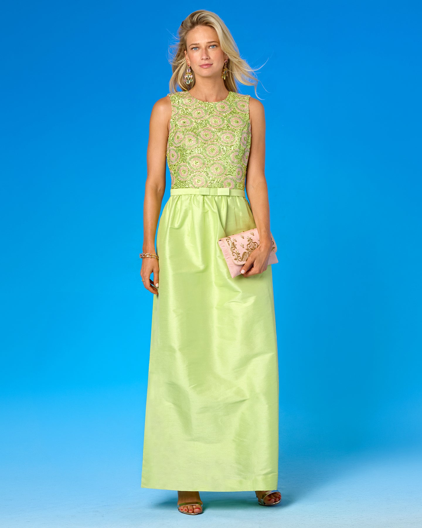 A woman modeling a sleeveless lime green maxi dress with a floral embroidered bodice and a satin-like skirt, holding a pink clutch against a solid blue studio background.