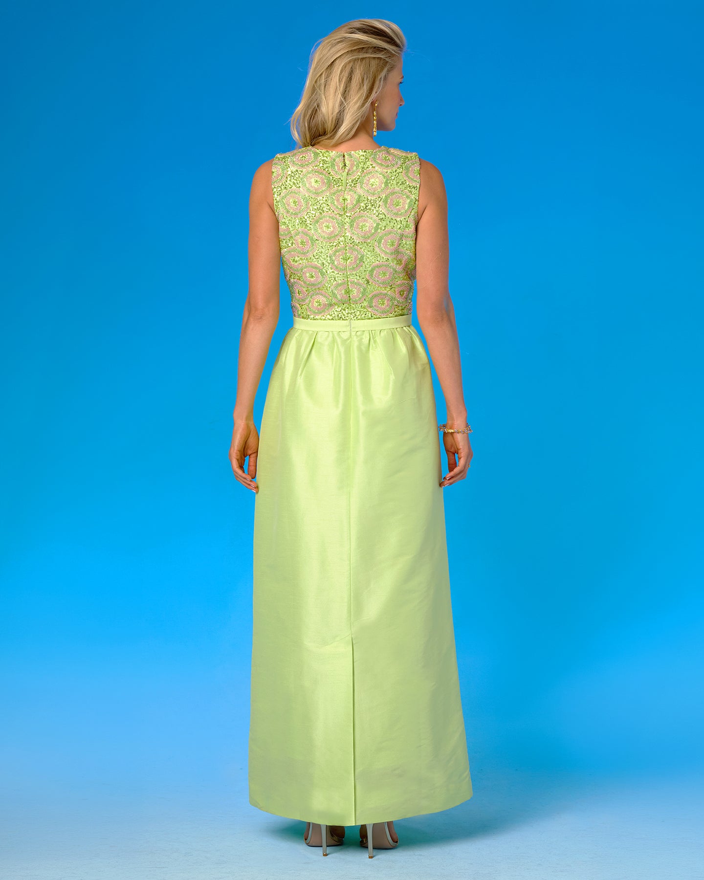 A woman viewed from the back wearing a sleeveless lime green formal gown with a patterned embroidered bodice and a solid satin skirt, set against a solid blue studio background.