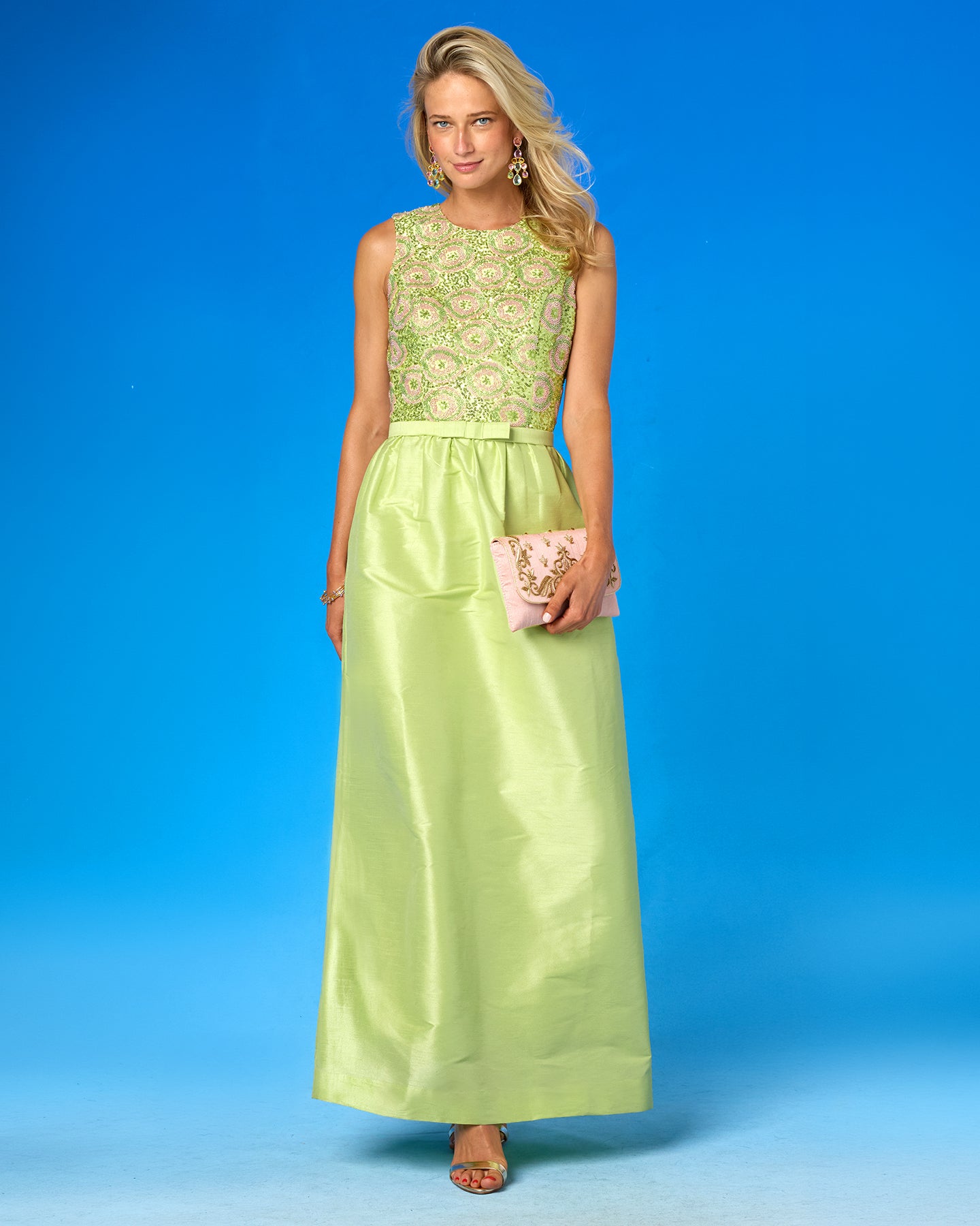 A woman models a sleeveless maxi dress with a light green and white floral lace bodice and a flowing lime green satin skirt, holding a small pink clutch against a bright blue studio background.