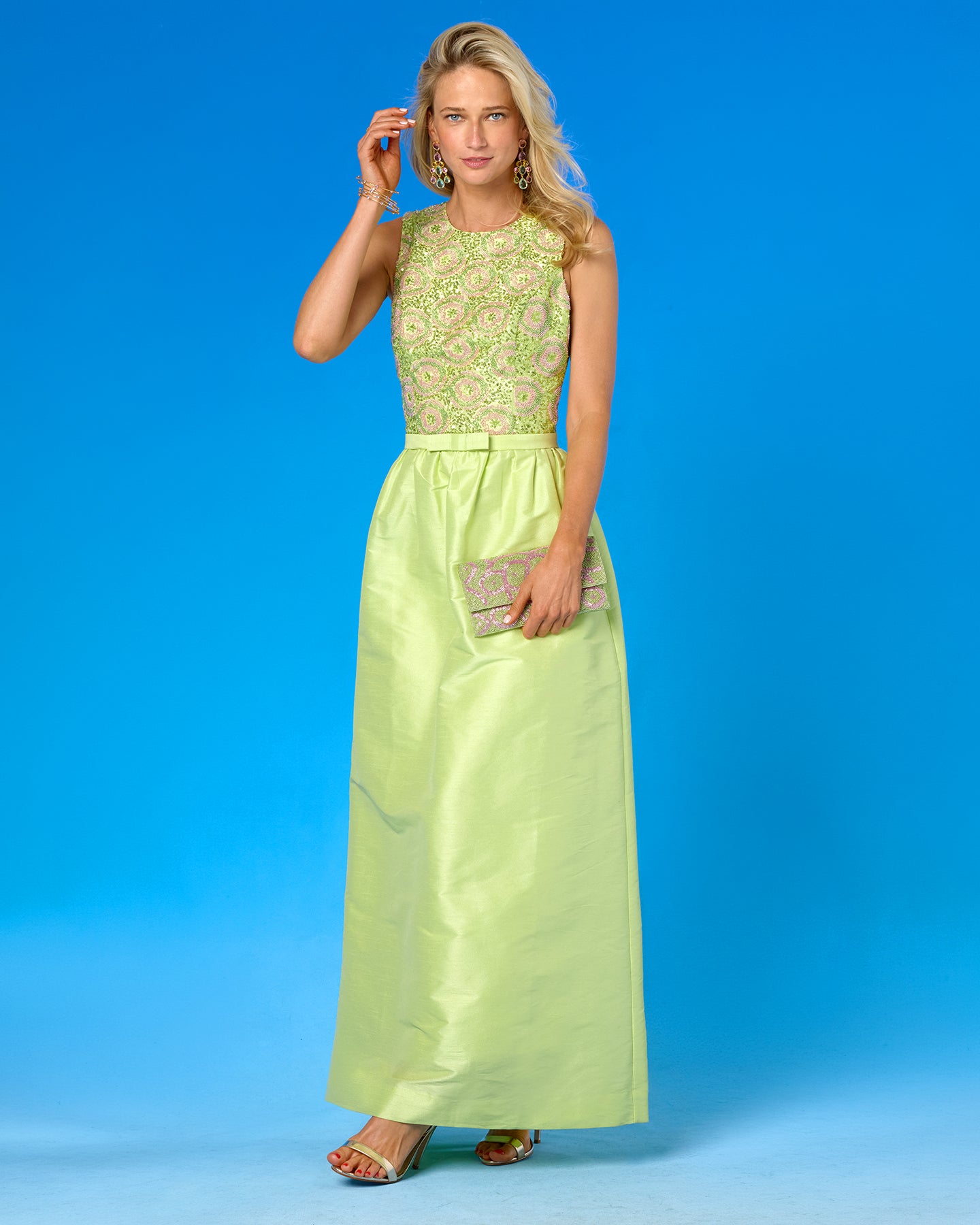 A woman modeling a sleeveless lime green maxi dress with a heavily embroidered bodice and a satin skirt, holding a metallic clutch against a solid blue studio background.