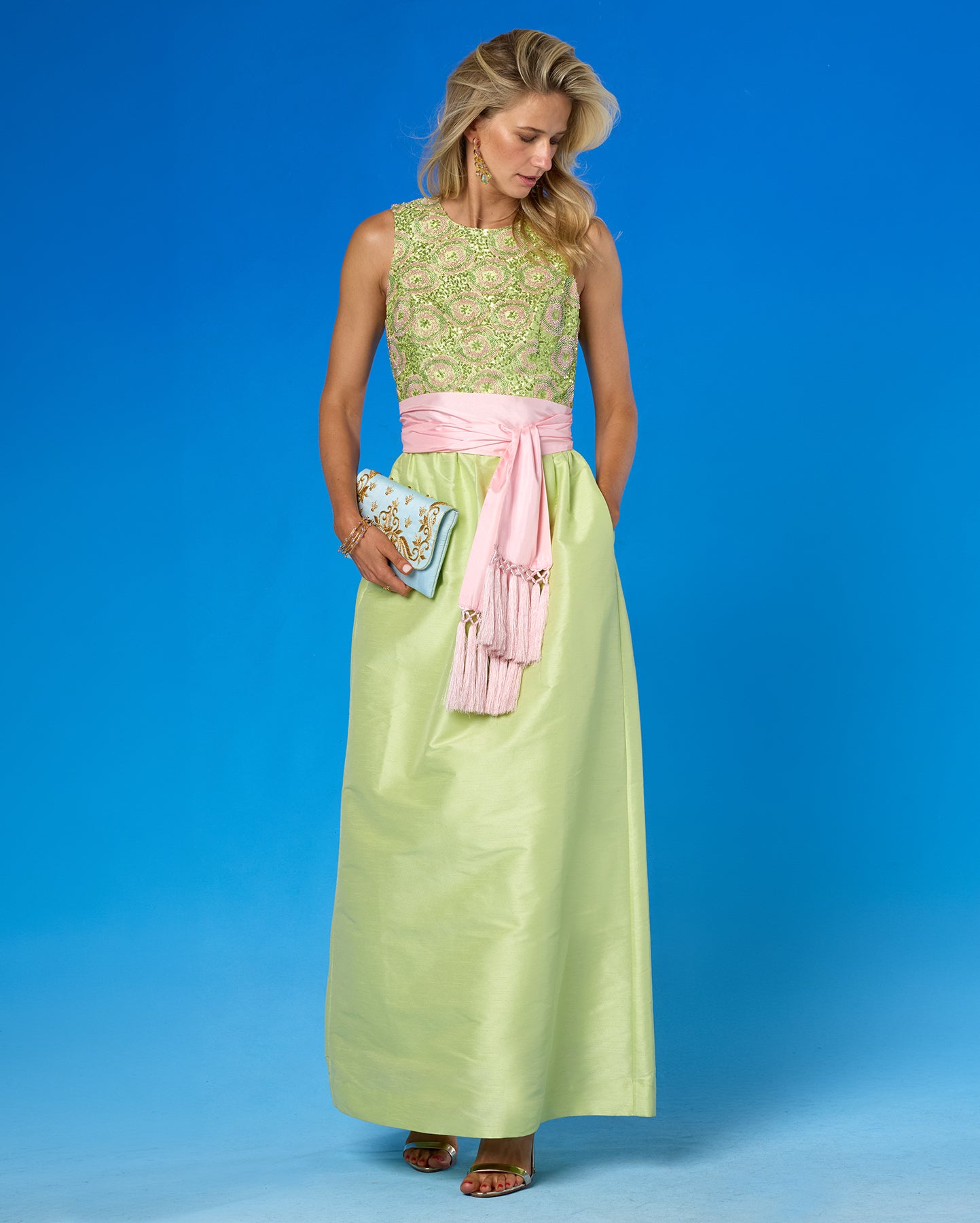 A woman modeling a sleeveless maxi dress with a light green embroidered bodice, a pink satin sash, and a light green satin skirt, standing against a blue studio background.