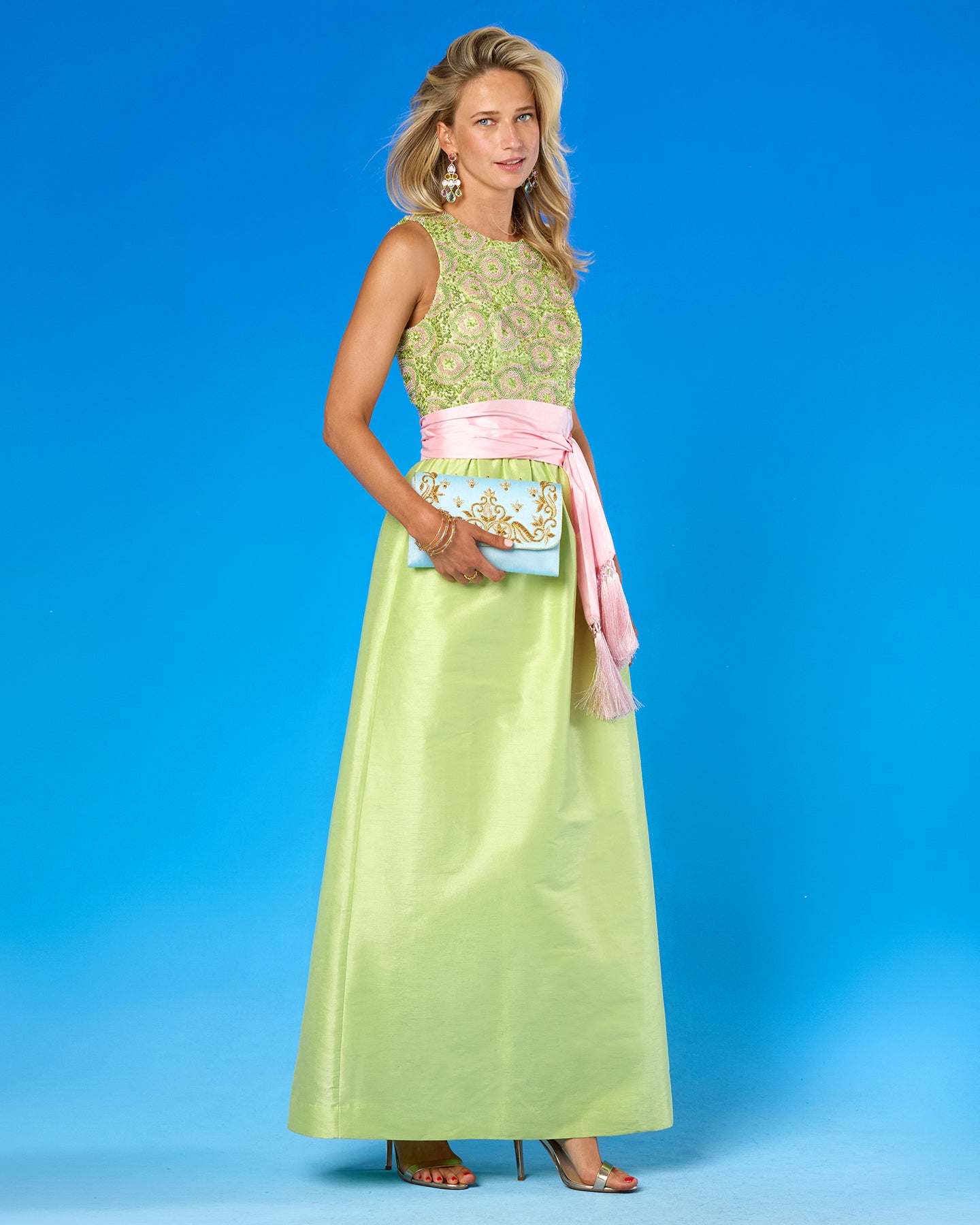 A woman models a sleeveless floor-length dress with a light green embroidered bodice, a pink sash, and a light blue clutch bag, set against a solid blue studio background.