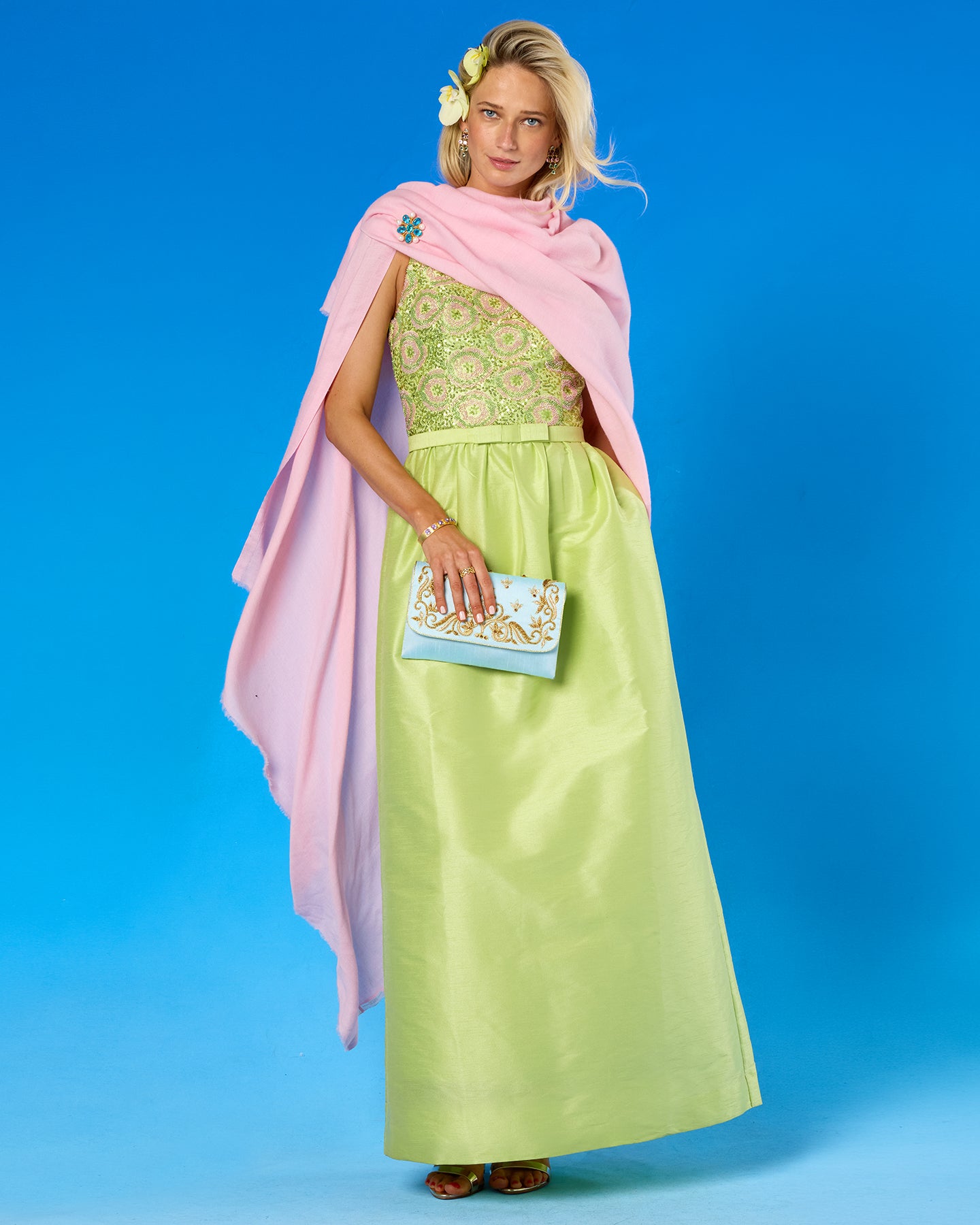 A woman models a long light green dress with a patterned bodice, a draped pink shawl, and a light blue clutch with gold embroidery, set against a solid blue studio background.
