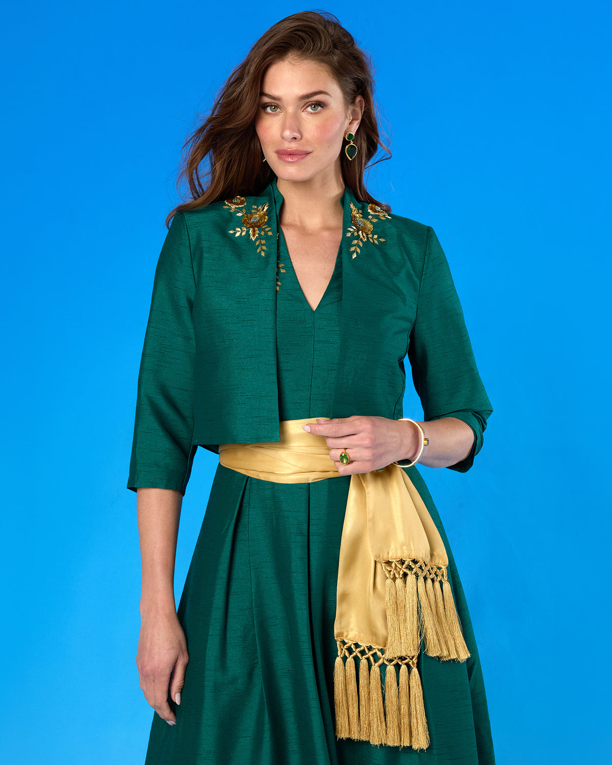 A woman modeling a green dress with a matching jacket, both decorated with gold floral embroidery, set against a solid blue background. She is holding a gold clutch and wearing a purple flower in her hair.