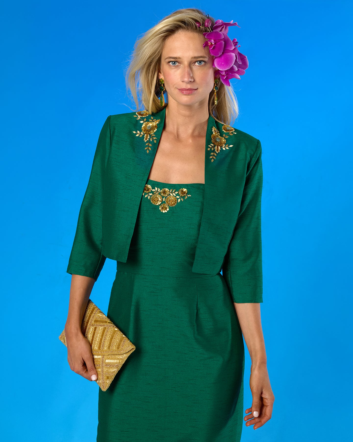 A woman models a dark green two-piece outfit consisting of a matching dress and cropped jacket, both decorated with gold floral embroidery. She is holding a gold clutch and wearing a purple flower in her hair, set against a solid blue background.