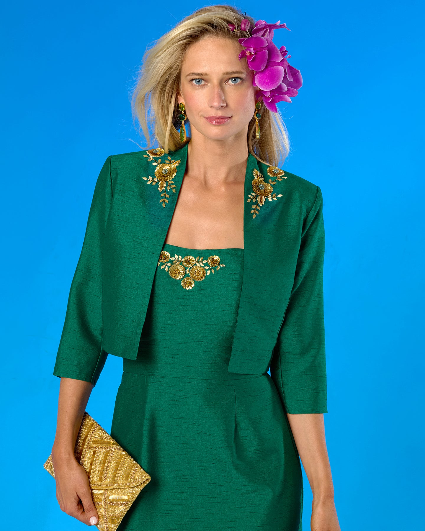 A woman modeling a green dress with a matching jacket, both decorated with gold floral embroidery, set against a solid blue background. She is holding a gold clutch and wearing a purple flower in her hair.