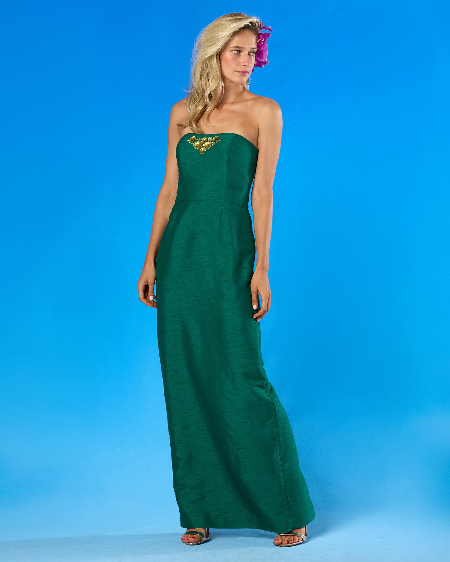 A woman modeling a strapless emerald green maxi dress with a gold embroidered chest detail, standing against a bright blue studio background.