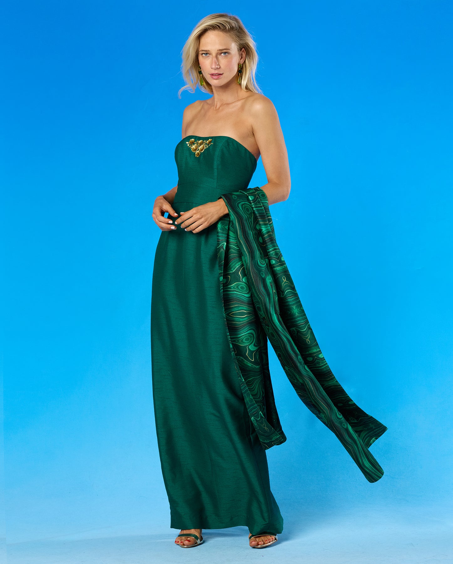 A woman modeling a strapless emerald green gown with a gold embellished bodice and a matching patterned green dupatta, set against a solid blue studio background.