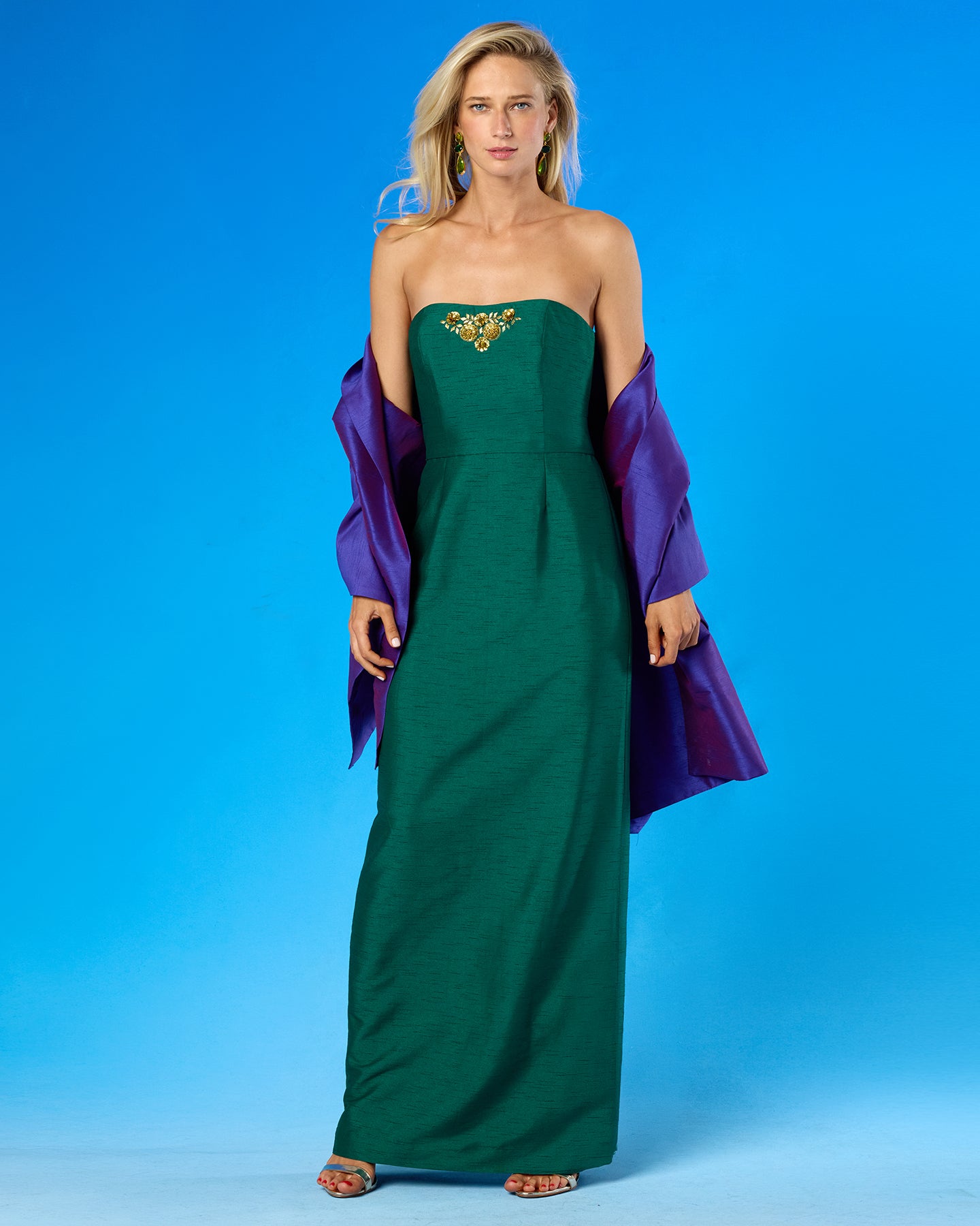 A woman models a strapless emerald green maxi dress with a gold embroidered neckline, styled with a draped purple shawl, against a solid blue studio background.