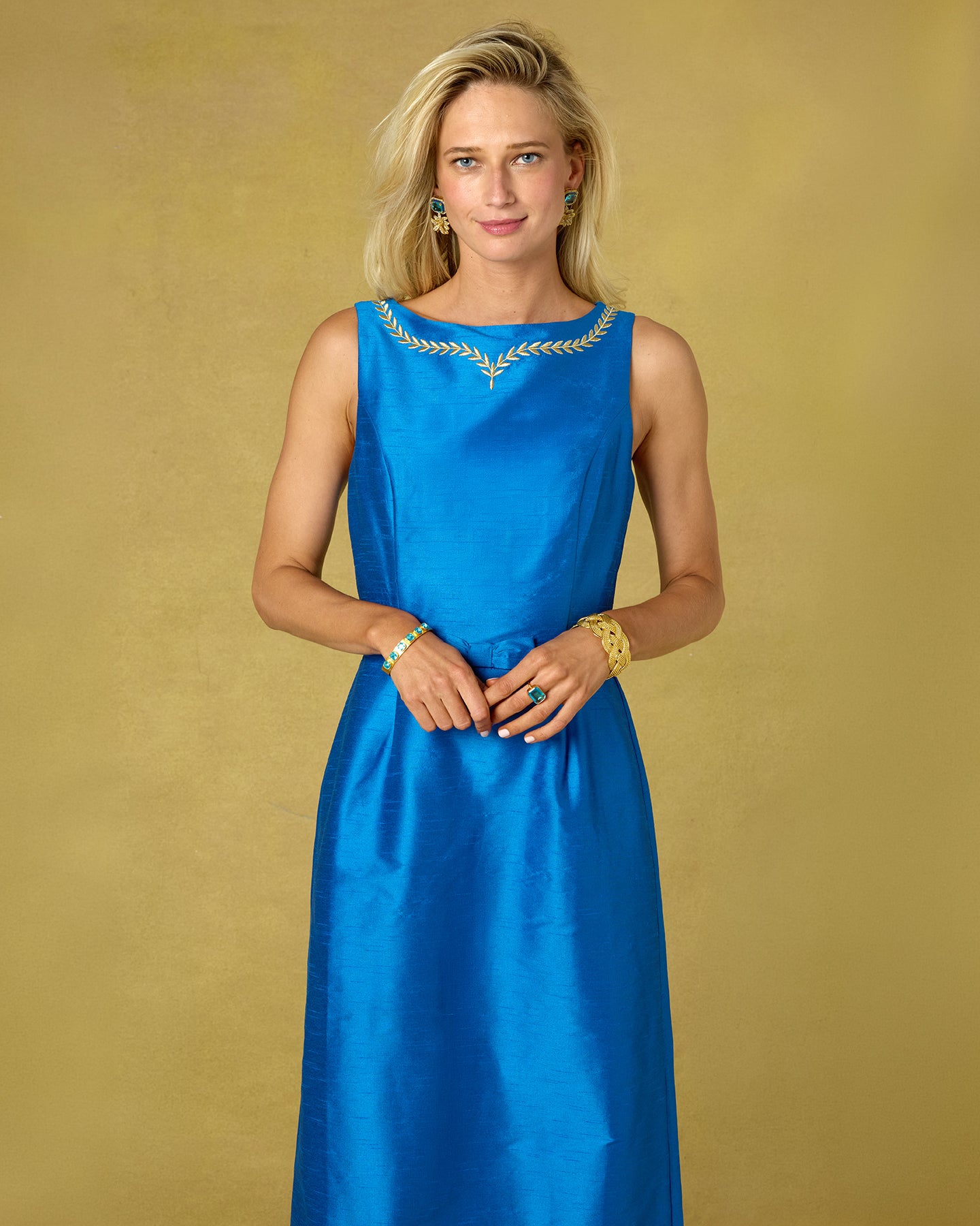 A woman modeling a sleeveless royal blue satin dress with gold embroidery around the neckline, standing against a textured gold background.