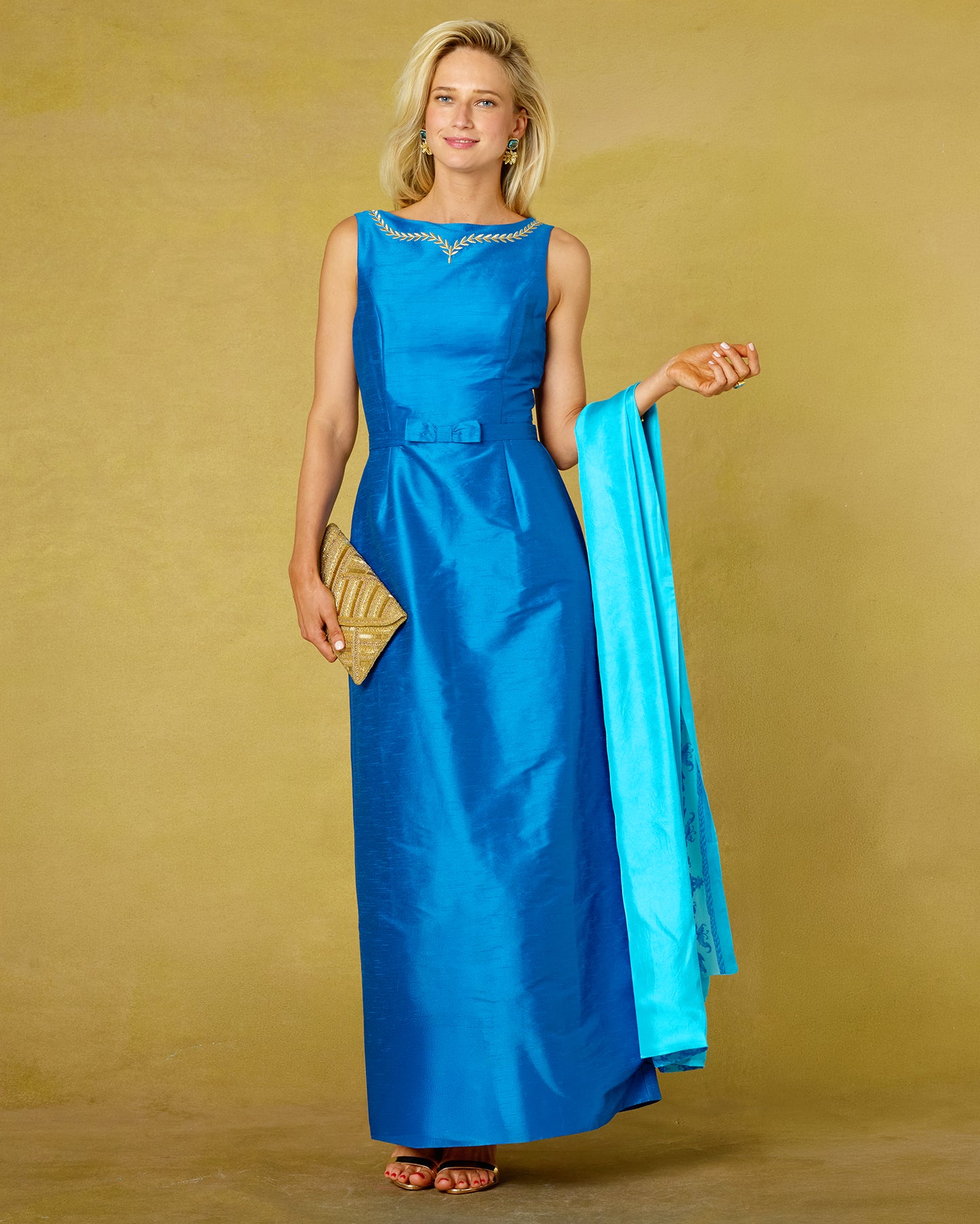A woman modeling a sleeveless bright blue floor-length gown with gold trim around the neckline, holding a matching blue fabric panel and a gold clutch against a warm tan studio backdrop.