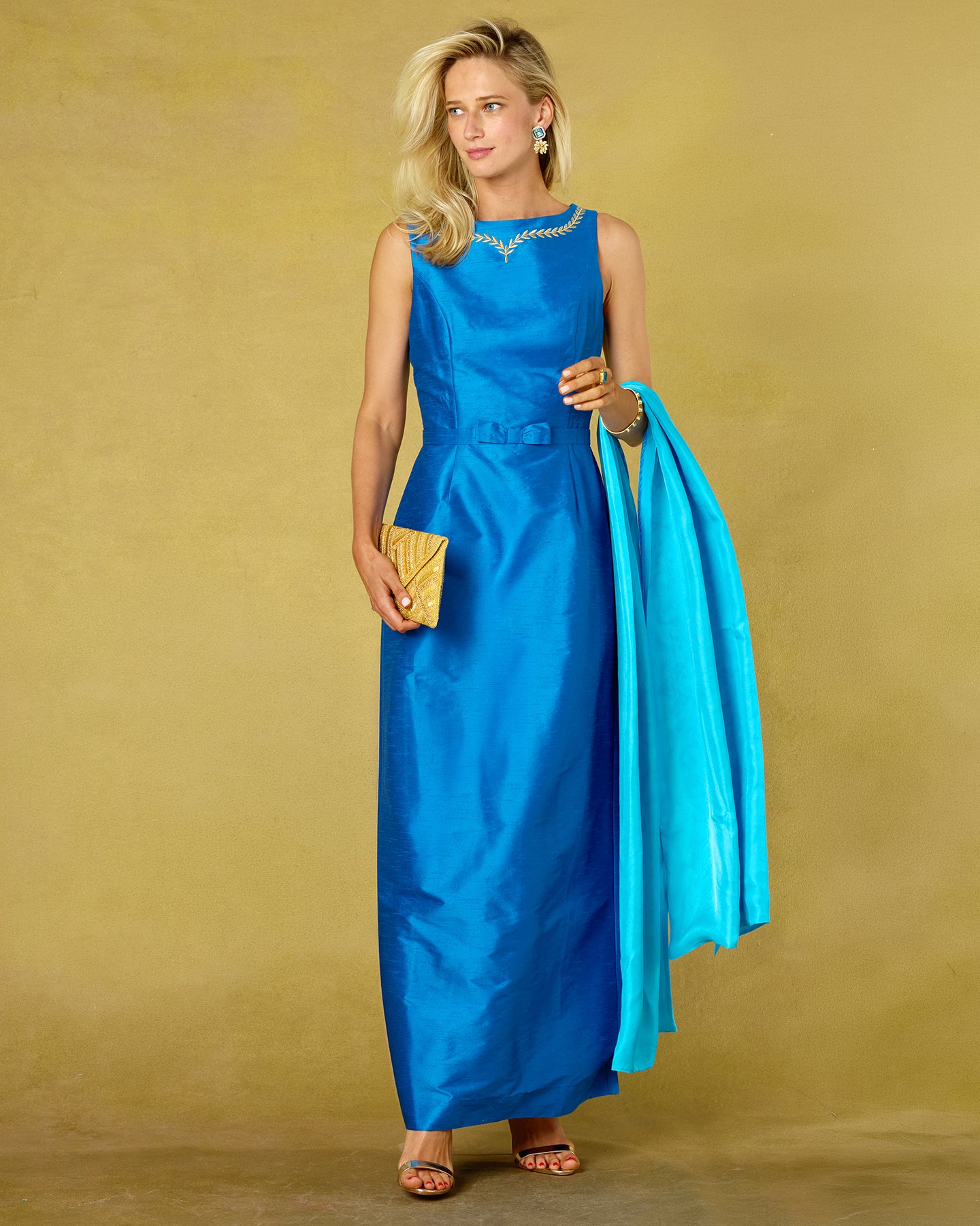 A woman models a sleeveless royal blue satin maxi dress with a matching belt, holding a bright turquoise dupatta and a gold clutch against a textured mustard yellow studio backdrop.
