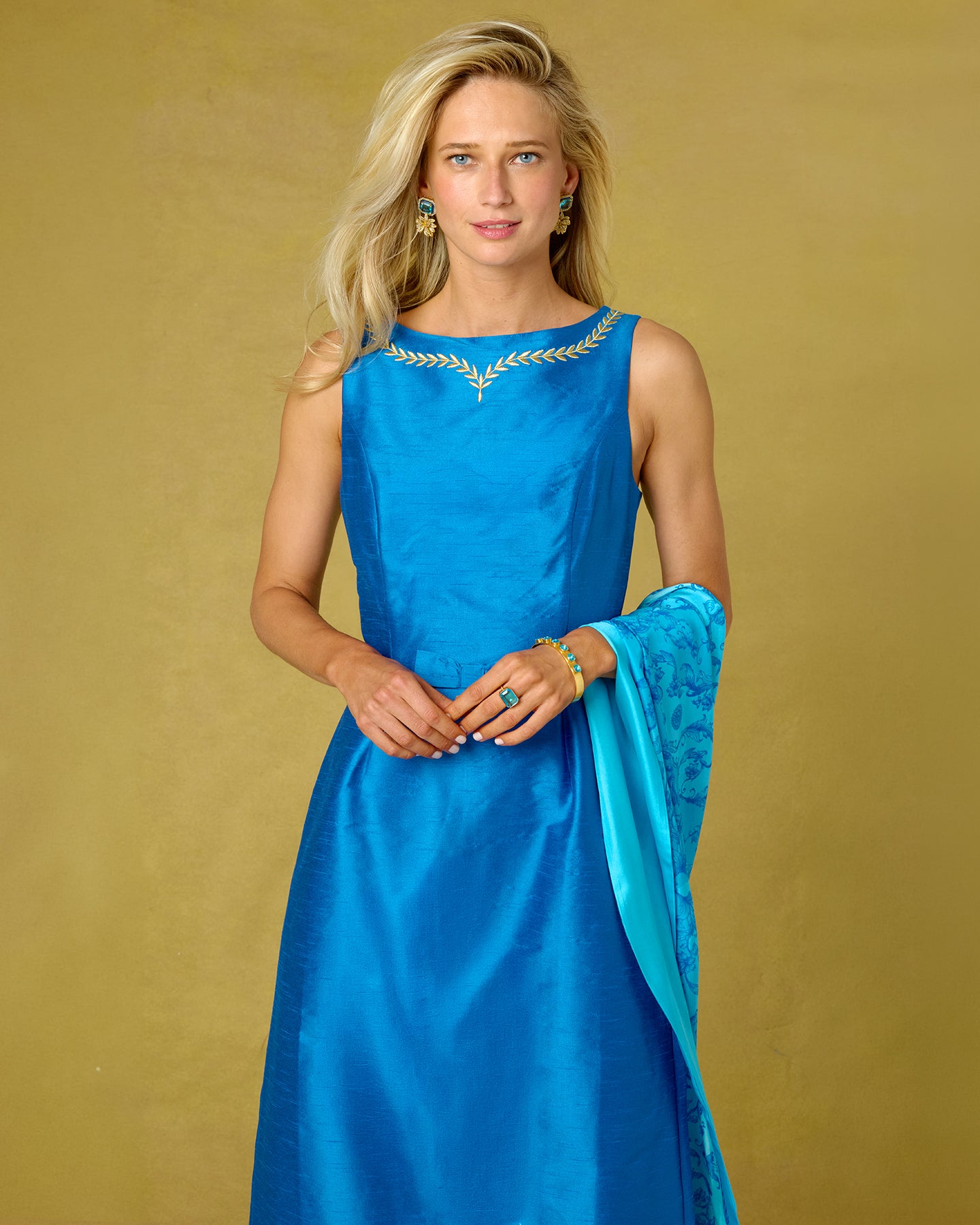 A woman modeling a sleeveless royal blue satin gown with gold leaf embroidery around the neckline, paired with a matching blue dupatta, set against a solid gold background.