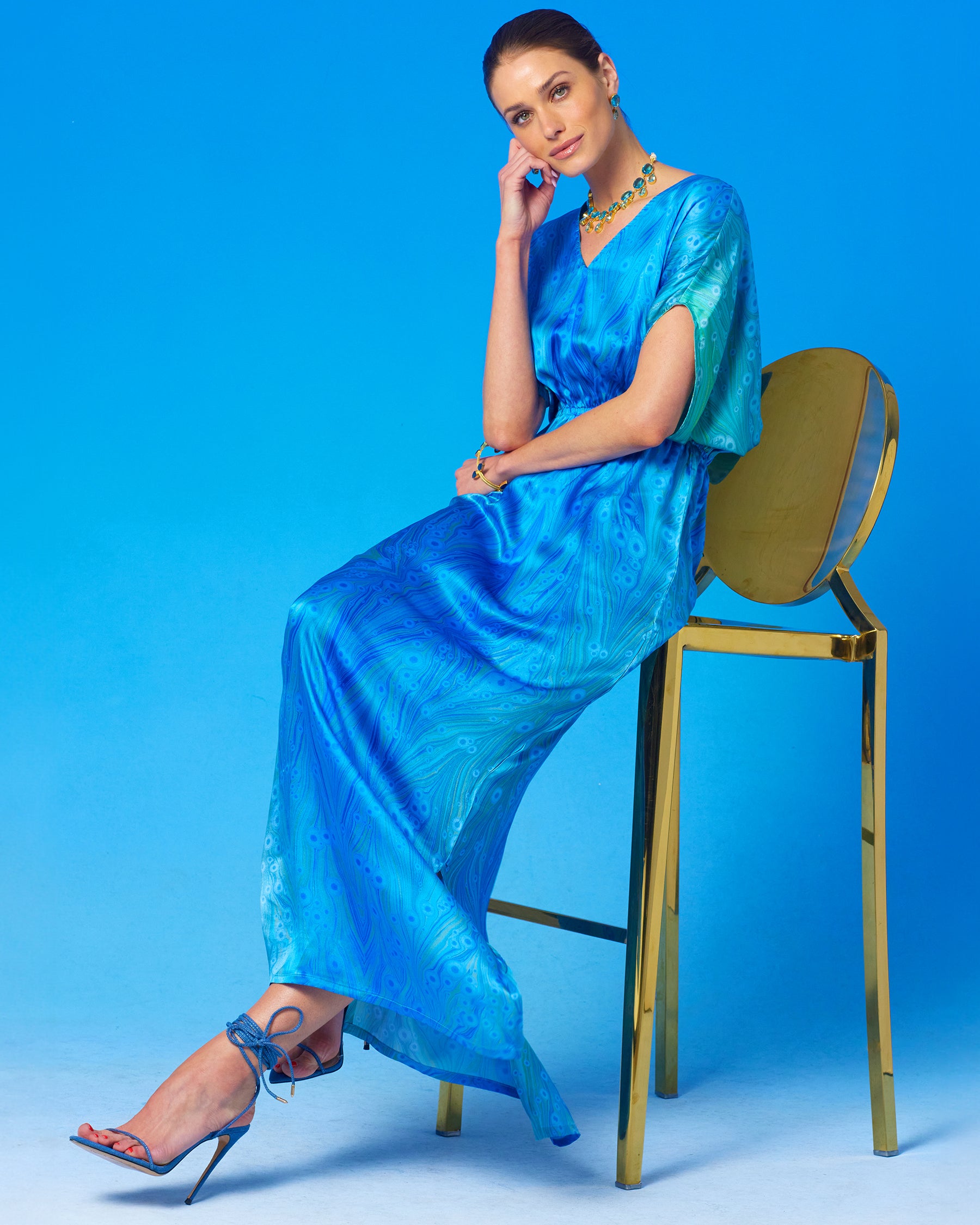 Calliope Long Silk Dress in Sea Nymph Blues witting on a gold chair