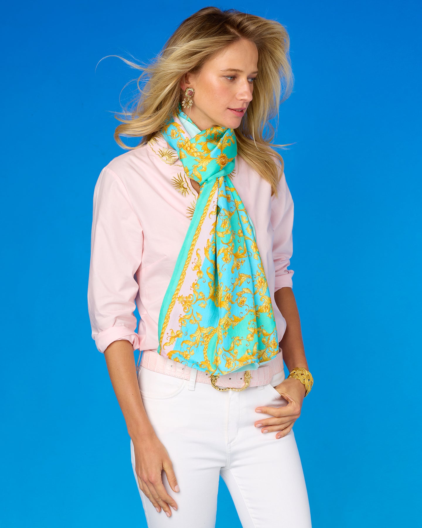 Woman wearing a pink shirt, white pants, and a turquoise and yellow patterned scarf against a blue background