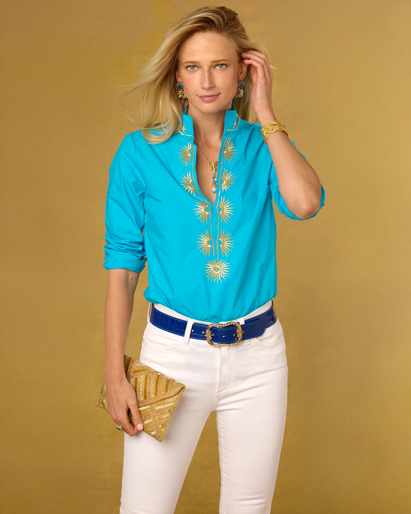 Woman wearing a turquoise blouse with yellow embroidery, white pants, and a gold clutch against a mustard yellow background.