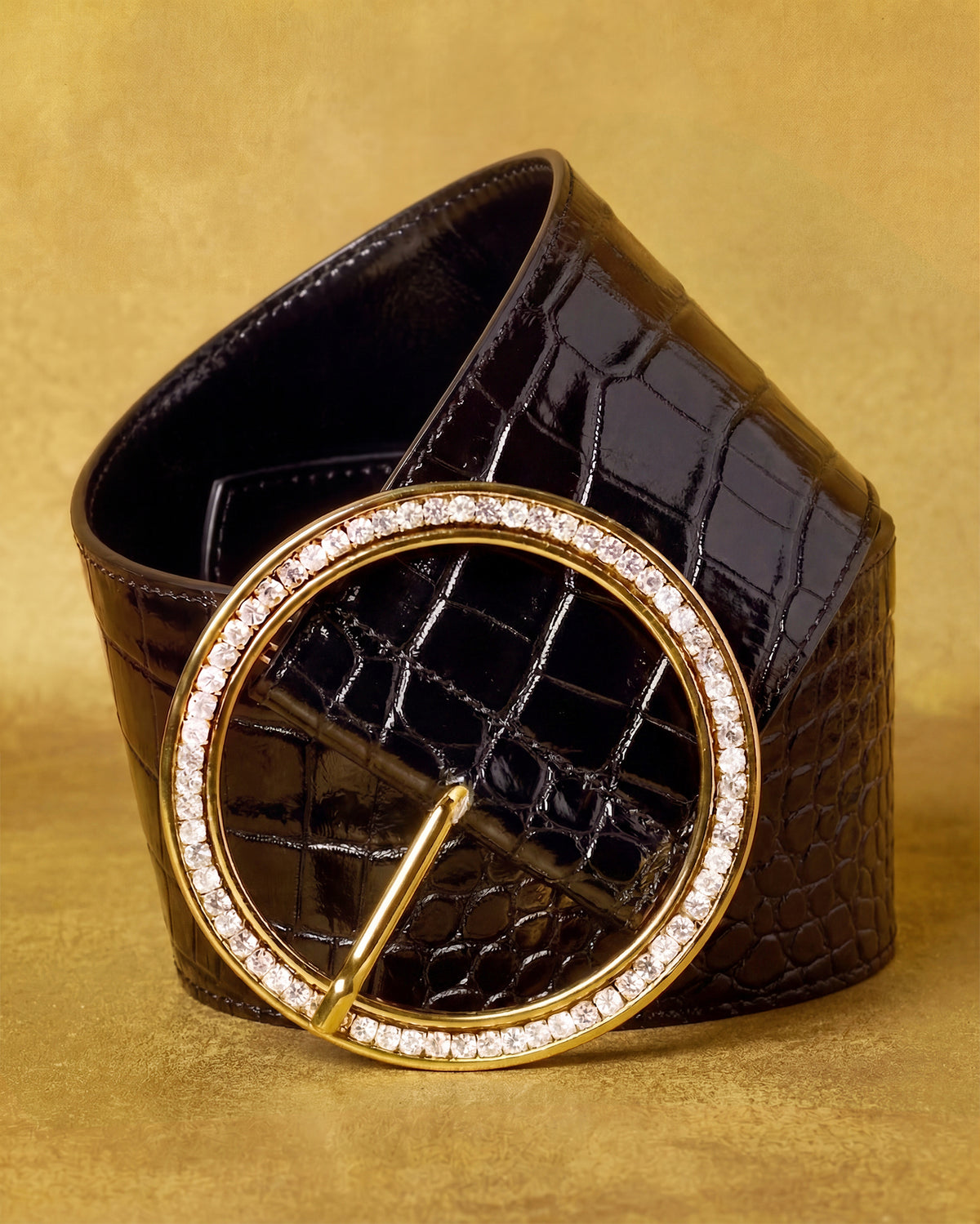 A dark brown crocodile-embossed leather belt with a large oval gold-tone buckle encrusted with clear rhinestones, displayed on a textured gold background.