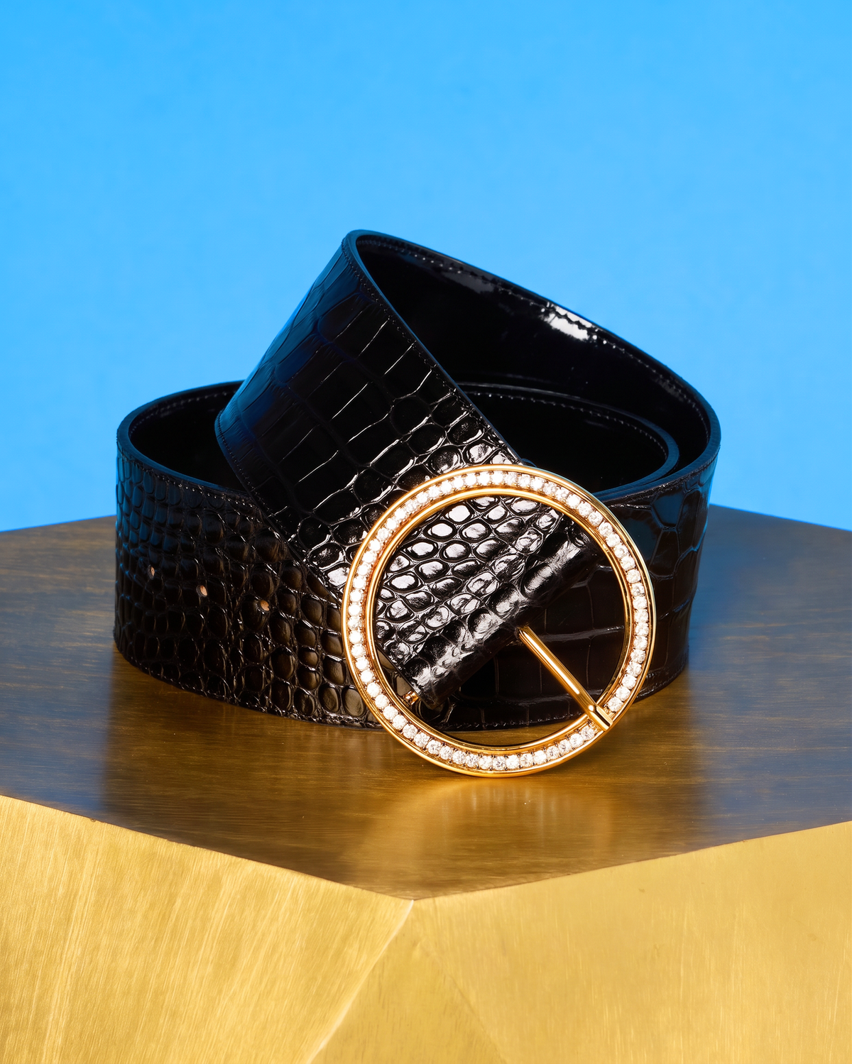 A black belt with a glossy crocodile-embossed texture and a large gold-tone circular buckle encrusted with clear rhinestones, displayed on a wooden surface against a bright blue background.