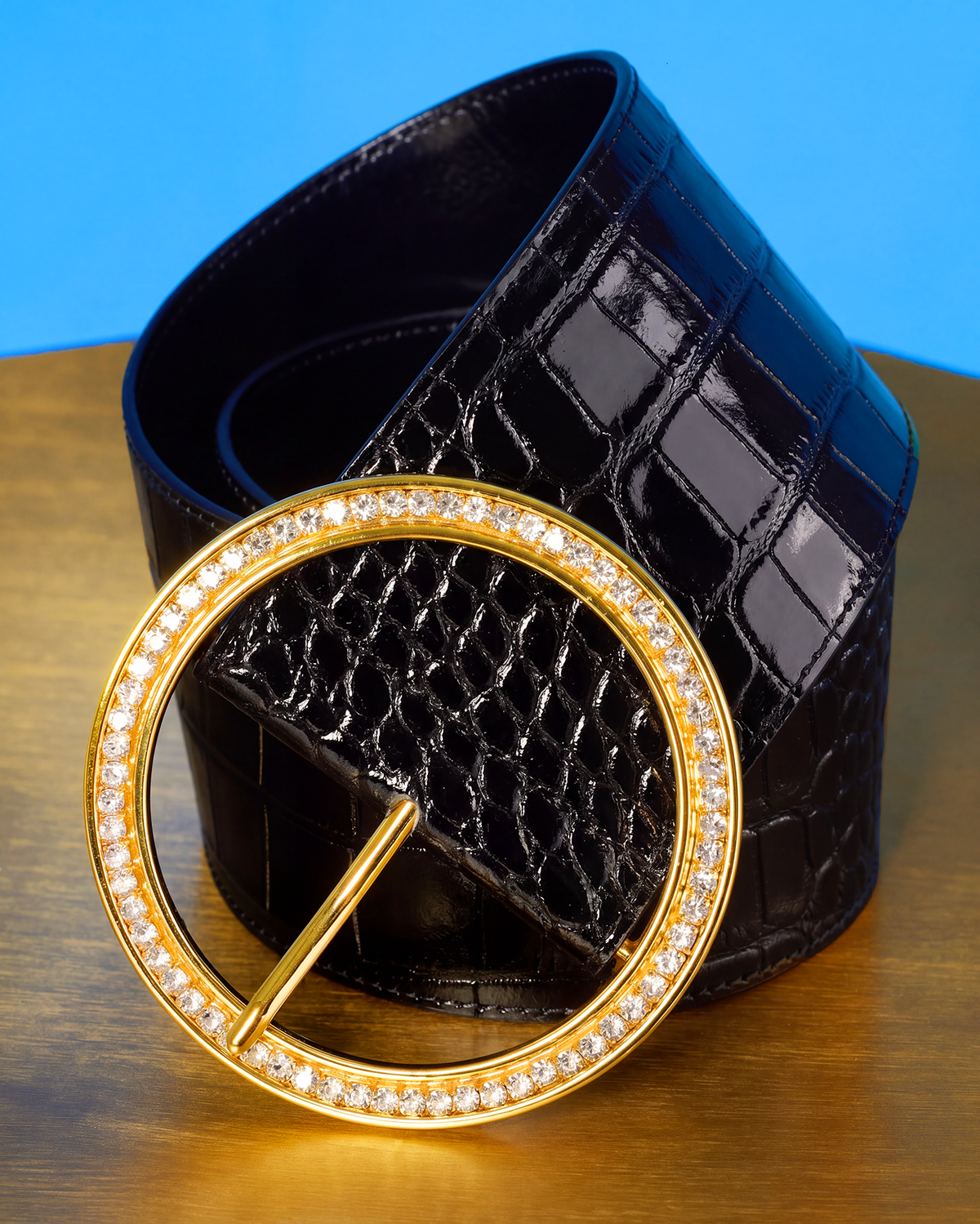 A black belt with a glossy crocodile-embossed texture and a large gold-tone circular buckle encrusted with clear rhinestones, displayed on a wooden surface against a bright blue background.
