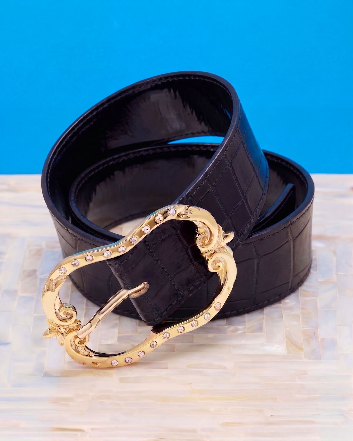 A black faux-crocodile leather belt with a large ornate gold-tone buckle adorned with small clear rhinestones, coiled on a light wood surface against a bright blue background.